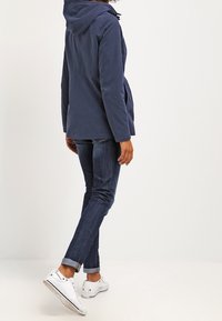 Mörkblå softshelljacka med huva, med avslappnad passform och knappdetaljer, bärs över mörka denimjeans och vita sneakers.