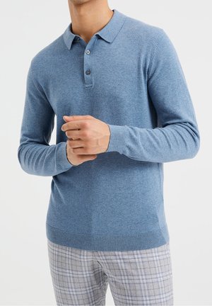 Man wearing a light blue long-sleeve polo sweater and gray plaid pants, hands clasped in front against a plain background.