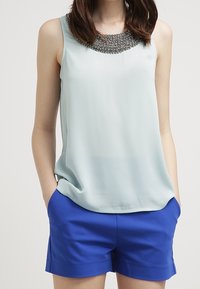 Top senza maniche azzurro chiaro con un collo decorato da perline, abbinato a pantaloni corti blu in un tessuto liscio con orli arrotolati.