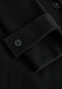 Melnā vilnas audumā ar mīkstu tekstūru, ar saliektu aproci un melniem divu caurumu pogām, kas izvietotas virsmā.