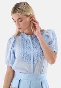 Hellblaue Bluse aus leichtem Stoff mit einer gerafften Vorderseite, kurzen Puffärmeln und einem Knopfverschluss.