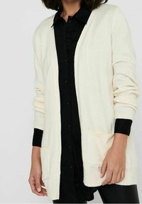 Ljus beige lång kofta i mjukt material med ribbade ärmslut, öppen front och framfickor, lager på lager över en svart skjorta.