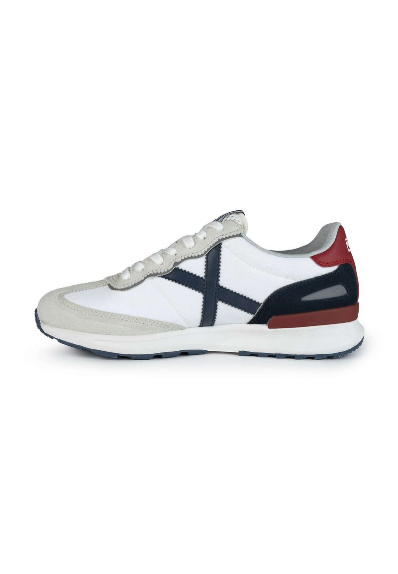Zapatilla deportiva con parte superior de textil blanco y acentos de ante gris, detalle de logo en azul marino, talón rojo y suela de goma con patrón de dibujo azul.