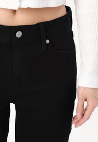 Κοντινό πλάνο ενός ατόμου που φορά μαύρα ψηλόμεσα τζιν και ένα λευκό ριμπ παιδικό άσπρο μπλουζάκι με μακρύ μανίκι, με το δεξί χέρι του χαλαρό στο πλάι.