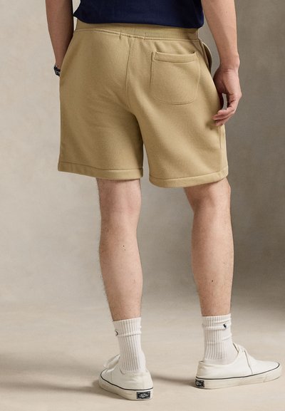 Shorts beige à texture douce, avec une poche arrière et des ourlets roulés, associés à des chaussettes blanches et des baskets sur un fond neutre.