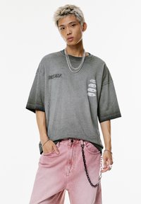 Ung person med afbleget hår iført oversized grå "TRUE SELF" t-shirt, lyserøde bukser med sort-og-sølv kæde, sølv halskæde og ringe.