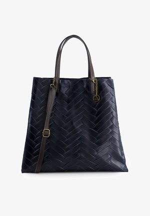 Granatowa skórzana torba typu tote z plecionym wzorem w jodełkę, dwoma ciemnobrązowymi uchwytami, regulowanym paskiem na ramię oraz małym złotym zawieszonym ozdobnym elementem.