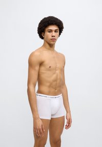 Λευκό βαμβακερό μποξεράκι με ελαστική μέση με λογότυπο, διακοσμημένη με μαύρες και κόκκινες λεπτομέρειες. Το μοντέλο στέκεται πλευρικά μπροστά σε απλό φόντο.