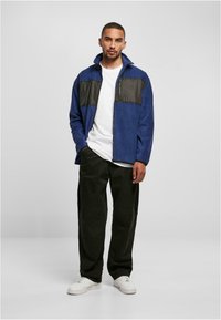 Giacca in fleece blu con pannelli neri, colletto alto, zip frontale, abbinata a pantaloni neri a gamba larga e sneakers bianche. Texture liscia.