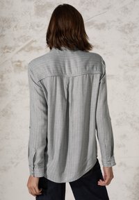 Lichtgrijze langslavige shirt met subtiele verticale strepen, knoopmanchetten en een ronde zoom, met een rugpas en plooien details.