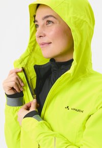 Vrouw in limoen groene hooded jacket, die het aantrekt, met zwart en grijs onderkleding zichtbaar en het VAUDE-logo op de borst van de jas.
