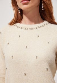 Pull en tricot crème orné de garnitures en argent et en perles, avec un col rond et une finition texturée et douce.