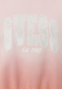 Felpa rosa chiaro con lettering perforato bianco di "GUESS" e "Est. 1981." Texture morbida con un effetto sfumato da un rosa chiaro a uno più scuro.