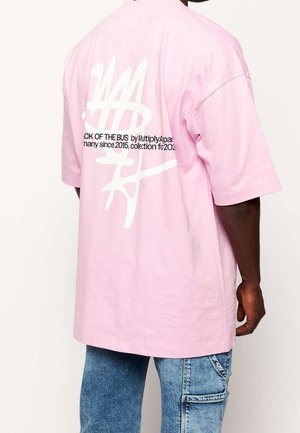 Man draagt een pastelroze oversized T-shirt met een witte abstracte afbeelding en gedeeltelijke zwarte tekst op de achterkant, gecombineerd met lichtblauwe spijkerbroek.