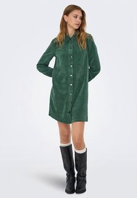 Grünes Cord-Hemdkleid mit Knopfleiste, Kragen, seitlichen Taschen und langen Ärmeln, kombiniert mit kniehohen schwarzen Stiefeln und weißen Socken.