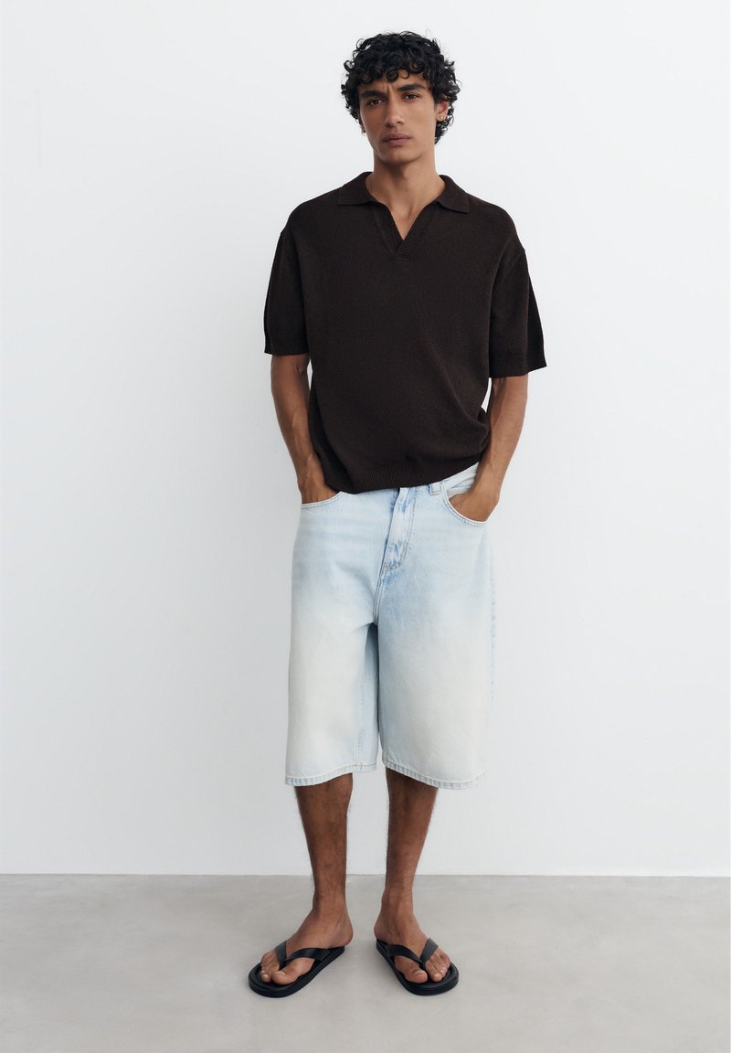 Homme aux cheveux bouclés portant un polo noir à manches courtes, un short long en denim clair et des tongs noires, debout les mains dans les poches.