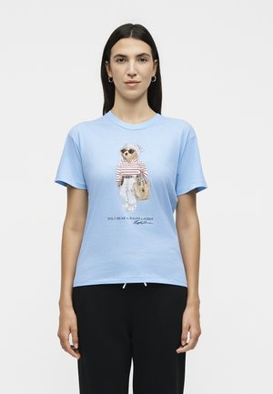 Γυναίκα με ίσια σκουρόχρωμα μαλλιά φορά ένα ανοιχτό μπλε T-shirt με μια αρκούδα με γυαλιά ηλίου, ριγέ τοπ, άσπρο παντελόνι και κρατά μια τσάντα tote.