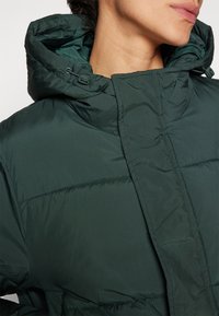 Veste rembourrée verte avec un col haut et une capuche réglable. Présente des coutures horizontales et une texture lisse. Détail de fermeture éclair visible.