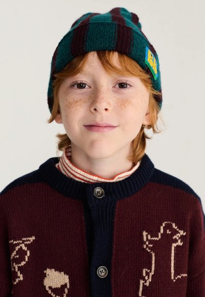 Jeune enfant roux avec des taches de rousseur portant un bonnet rayé vert et marron et un cardigan bordeaux avec des motifs d'animaux.