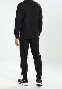 Czarna bluza z długim rękawem i dresowe spodnie z białymi paskami po bokach. Wyposażone w okrągły dekolt i ściągacze na rękawach. Noszone z czarnymi butami sportowymi.