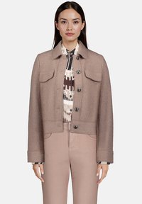 Femme portant une veste brune cintrée avec des boutons sur une chemise à motifs et un pantalon marron assorti, se tenant devant un fond uni.