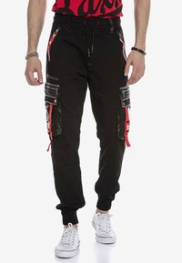 Schwarze Cargo-Jogginghose mit mehreren Reißverschlusstaschen, roten Akzenten und elastischen Bündchen. Verfügt über ein glänzendes schwarzes Panel und eine strukturierte Oberfläche.
