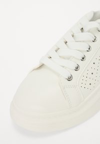 Hvit lær sneakers med perforerte detaljer, flate hvite lisser og sølvfargede øyer, som har rund tå og en teksturert gummisåle.