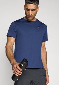 Μπλουζάκι αθλητικό σε ναυτικό μπλε με κοντά μανίκια και ασημί λογότυπο. Ένα άτομο κρατά ένα μαύρο μπουκάλι νερού με καπάκι βιδωτό.