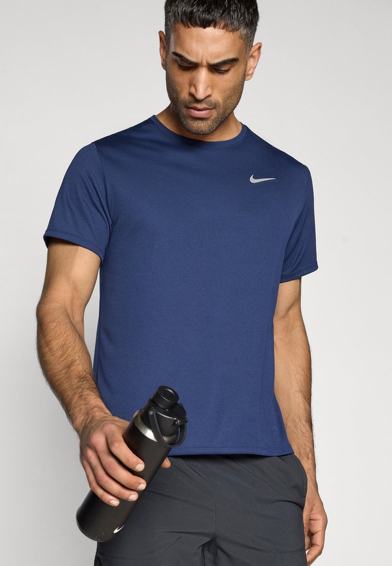 Marineblaue Sport-T-Shirt mit kurzen Ärmeln und silbernem Logo. Eine Person hält eine schwarze Wasserflasche mit Schraubverschluss.