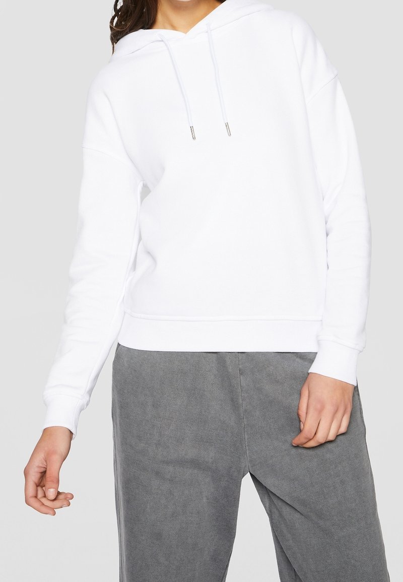 Personne portant un sweat à capuche blanc uni avec cordons et un pantalon gris ample, debout devant un fond gris neutre.