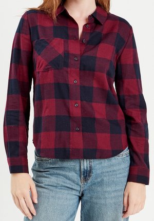 Femme portant une chemise à carreaux rouge et noir boutonnée avec manches longues et un jean bleu clair sur un fond uni.
