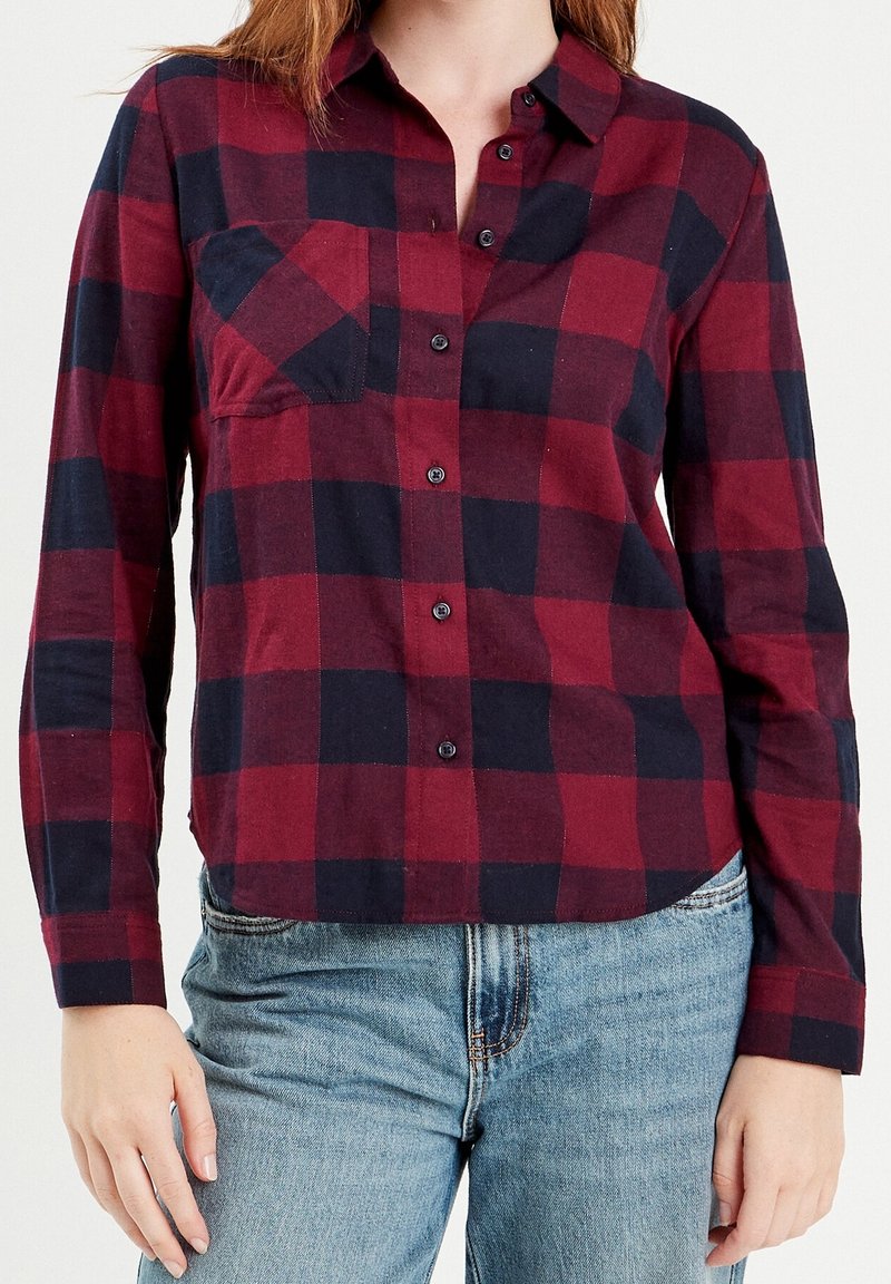 Femme portant une chemise à carreaux rouge et noir boutonnée avec manches longues et un jean bleu clair sur un fond uni.