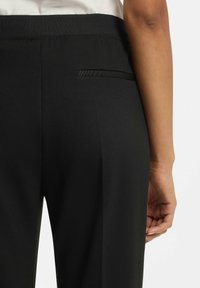 Sorte bukser med glat stof, elastisk linning og en enkelt baglomme med slank design. Nærbillede af bagfra.