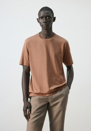 Mladý muž stojaci s jednou rukou v kapse, oblečený v lososovo sfarbenom tričku a béžových nohaviciach na jednoduchom svetlom pozadí.