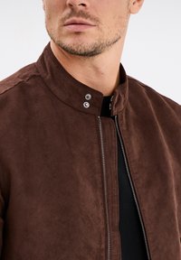 Bruine leren jas van suede met een staande kraag, voorrits en drukknopen als accenten. Zachte textuur en slank ontwerp.