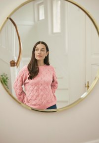 Rosa gestrickter Pullover mit Zopfmuster, langen Ärmeln und Rundhalsausschnitt, kombiniert mit blauen Jeans; reflektiert in einem runden Spiegel mit goldenem Rahmen.