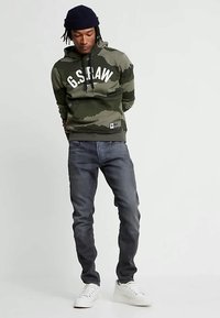 Muškarac nosi kapu s uzorkom zelene vojske G.S. RAW, tamne traperice, bijele tenisice i crnu kapu, stojeći s rukama iza leđa.