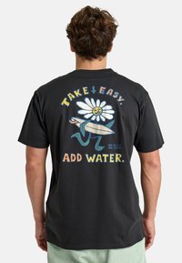 T-shirt en coton noir avec un graphisme coloré représentant une fleur souriante, une planche de surf et le texte "PRENDS LES CHOSES FACILE. AJOUTE DE L'EAU." au dos.