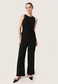 Top sin mangas de canalé negro, combinado con pantalones anchos de encaje texturizado con patrón floral. La modelo lleva tacones negros de punta. Fondo neutro.