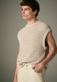Pull sans manches en maille beige léger avec un motif texturé en losanges. Il présente un col rond et des bords bruts. Associé à un pantalon léger.