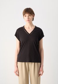 T-shirt nera a V con maniche corte, realizzata in tessuto morbido. Presenta un sottile bordo testurizzato lungo il colletto, abbinato a pantaloni beige.