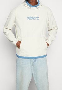 Ljus beige, huvad vindjacka med blå detaljer och "adidas ADVENTURE"-logotyp. Elastiska muddar, känguruficka och ett lager med blå innerfoder.