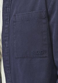 Camisa azul marino de tejido texturizado con un bolsillo en el pecho y detalle bordado "R.D.D.". Pliegue de botones visible en el lateral.