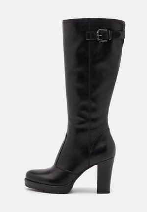 Botte en cuir noire hauteur genou avec un talon épais et haut, et une sangle à boucle ajustable près du sommet.