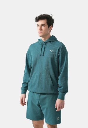 Junger Mann trägt einen türkisfarbenen Puma-Hoodie und passende Shorts, steht vor einem schlichten hellen Hintergrund und schaut zur Seite.