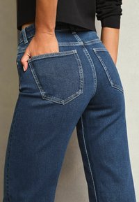 Temno modri jeans s hrbtnimi žepi, s ravnimi hlačnicami in kontrastnim šivanjem. Tkanina ima teksturiran videz.
