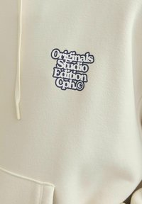 Felpa color crema con cappuccio, dotata di un cordino e un grande logo stampato in blu navy: "Originals Studio Edition Cph©" sul davanti.