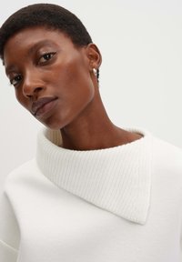 Maglione bianco lavorato a maglia con colletto largo a coste. Texture liscia e taglio dritto. Il modello mostra la parte superiore su uno sfondo neutro.