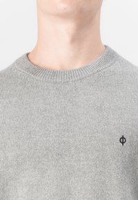 Grå stickad tröja med ribbad halsringning och svart logotypdetalj på bröstet. Mjuk textur, klassisk rund design och figurnära stil.