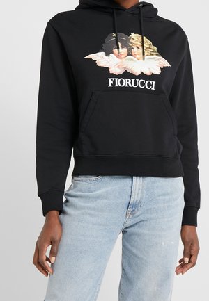 Personne portant un sweat à capuche noir Fiorucci avec un motif de jumeaux anges et un jean bleu clair sur un fond clair uni.